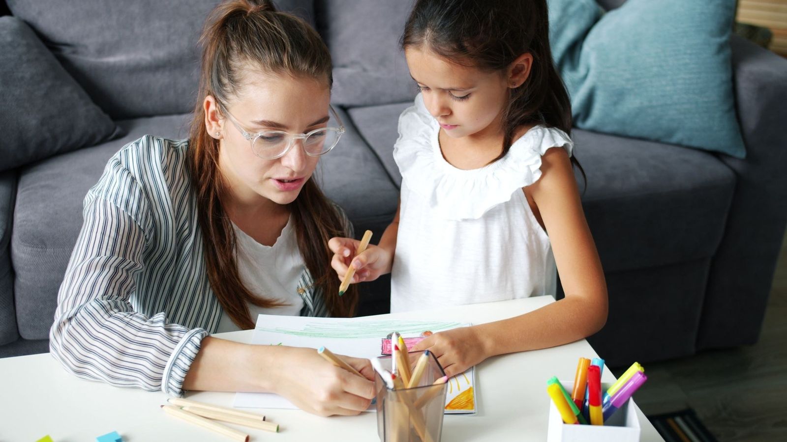 A girl and her tutor working with pencils at a table, focusing on learning.